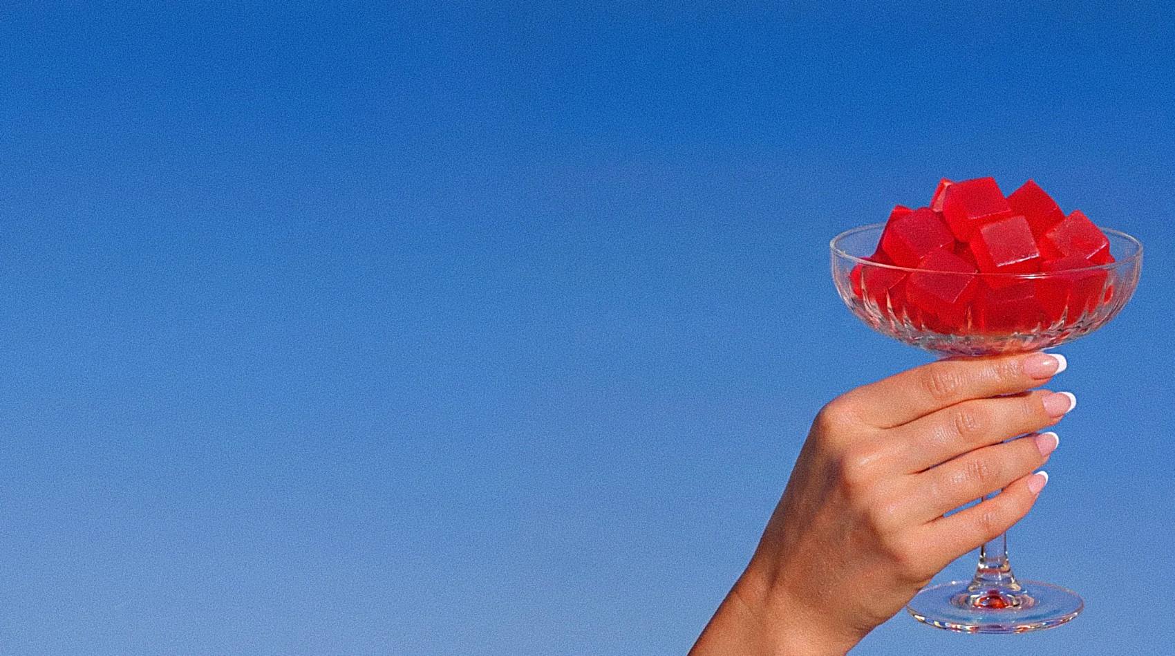 A hand raising a coupe glass piled with red Reform electrolyte gummies against a vivid blue sky